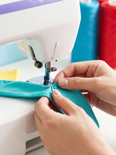 Close-up of hands carefully stitching a turquoise pet bandana on a sewing machine, colorful fabric rolls in the background, bright natural daylight