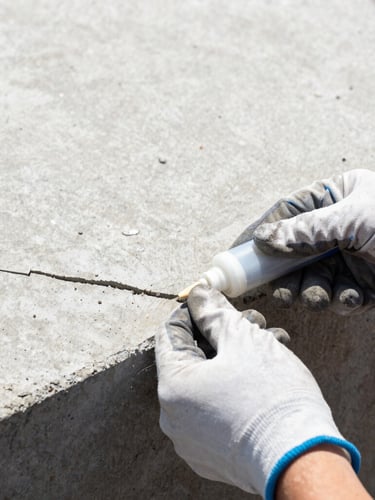 Close-up of a technician's gloved hands sealing a small crack in a concrete foundation with sealant, bright daylight, focus on the hands and crack.