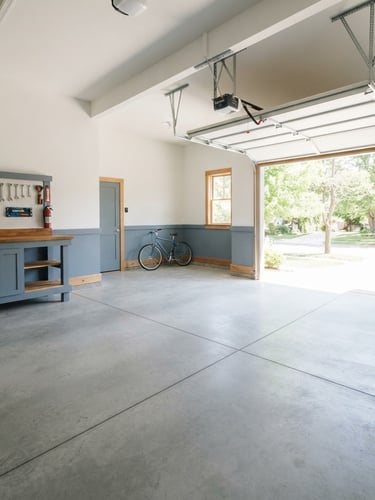 Wide-angle clean documentary photograph of a spacious, empty garage with pristine concrete floors, bright natural daylight streaming through the open door, slate blue accents