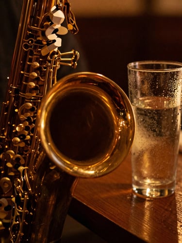 Close-up of a brass saxophone bell reflecting warm golden stage lights, with a condensation-rimmed highball glass sitting on a dark wooden table in the soft-focus background.