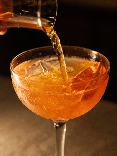 Close-up of a vibrant, garnished cocktail being poured, warm low-key evening light with rich golden accents, shallow depth of field, 35mm film.