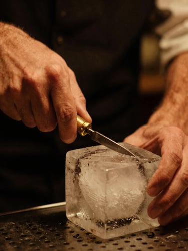 Close-up of a bartender carving a clear block of ice with a brass tool, warm low-key amber lighting, rich saturated tones, 35mm film