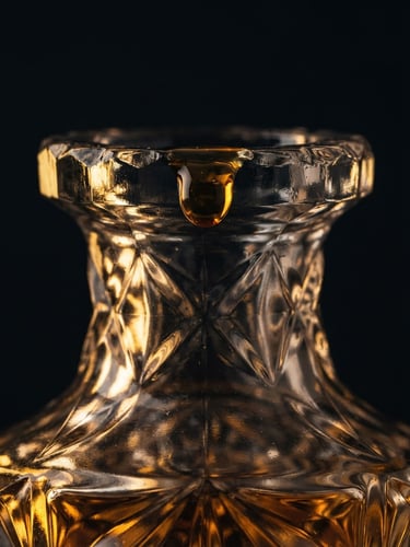 Macro portrait of a heavy crystal flacon reflecting golden side-lighting, amber oil droplet suspended on the rim, matte black background