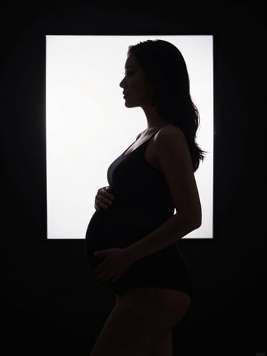 Dramatic maternity silhouette framed in absolute black, cinematic backlighting, asymmetric composition.