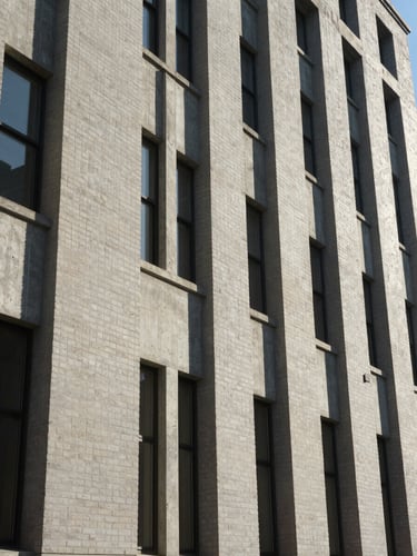 Wide environmental shot looking up a commercial building facade from street level, natural daylight raking across concrete and brick texture, strong vertical lines, no people, cool neutral light revealing material condition