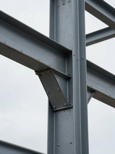 Close architectural detail of a commercial building's structural steel beam connection, natural overcast daylight raking across riveted metal plate and bolt heads from the left, shallow depth showing the joint's material weight and fabrication specificity, framed tightly to fill the vertical column