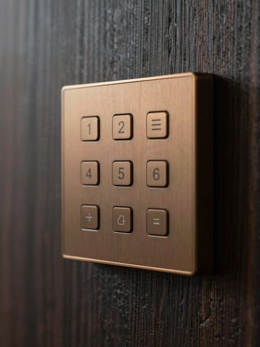 Close-up of a sleek, minimalist bronze home automation keypad mounted on a dark textured wood wall panel, soft warm architectural glow highlighting the clean lines, shallow depth of field.