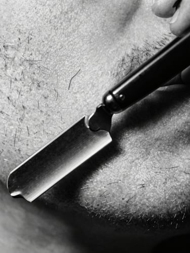 Macro extreme close-up of a straight razor carving a sharp line on a beard, high-contrast black and white, side-lit studio lighting, dramatic focus on the steel blade