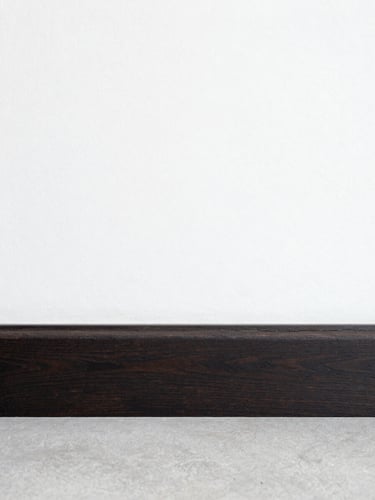 Straight-on architectural shot of a perfectly aligned, hand-wiped dark wood baseboard meeting a crisp linen-white wall, bright natural daylight, clean lines.
