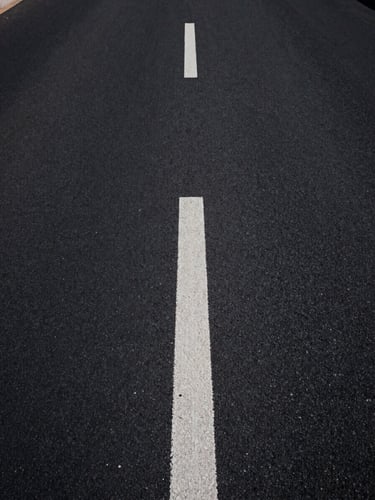Extreme close-up overhead view of a clean asphalt road stretching into the distance, a single white dashed center line cutting through deep shadow and pale light, minimal and geometric, abstract road geometry with no people, high contrast studio-quality lighting, wide negative space on the left side