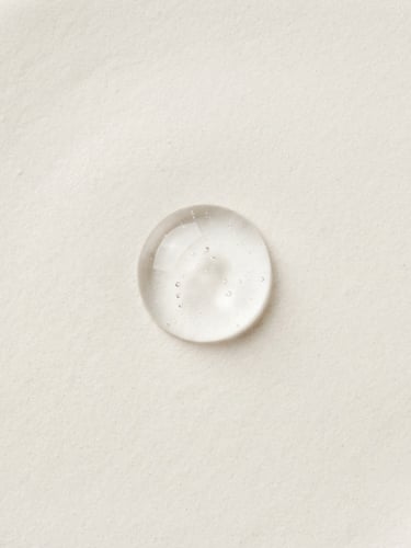 Overhead macro photography of a single clear cosmetic gel droplet on a warm off-white surface, bright diffused clinical studio light, sharp focus on texture