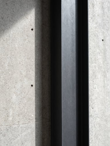 Macro photograph of a raw concrete wall meeting a clean black steel structural column under harsh directional sunlight, highlighting textures.