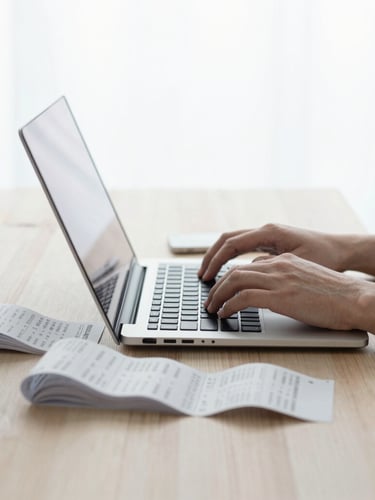 Hands typing on a laptop next to organized paper receipts on a light wooden table, bright overcast daylight, minimalist workspace