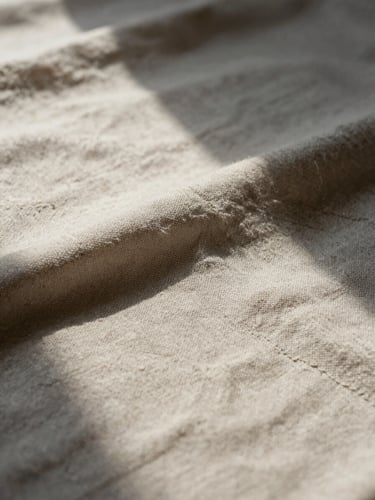 Extreme close-up of a finished textile piece laid on a raw linen work surface, intricate stitching visible mid-frame, soft north-facing daylight raking across the texture from the left, deep shadow in the folds, no props