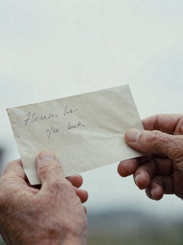 Close-up of aged hands gently holding a faded, handwritten letter, soft overcast daylight, desaturated tricolor tones, deep shadows, 35mm film grain