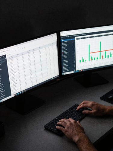 Wide overhead shot of a dark desk with two monitors displaying campaign performance dashboards, budget allocation tables and ROI delta charts visible, a hand resting near a trackpad, cool studio lighting, high contrast, no face visible