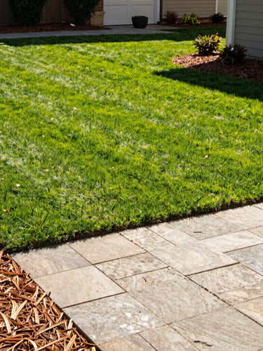 Bright outdoor photography of a pristine residential backyard with freshly cut green lawn, clean-edged mulch beds, and a stone paver patio under warm afternoon sunlight, 35mm