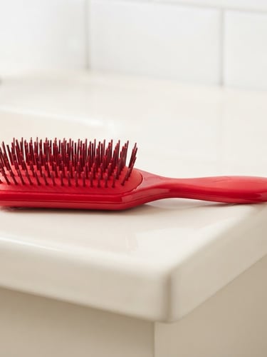 High-contrast product shot of a vibrant red curl-defining brush resting on a clean cream-colored bathroom shelf, bright high-key studio light, sharp focus, 35mm