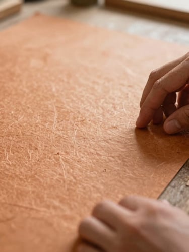 Close-up of hands gently inspecting the texture of thick, handmade paper under soft workshop light, with visible fibers and natural imperfections, shallow depth of field, warm editorial photography.