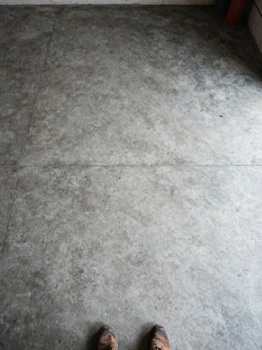 Overhead wide-angle shot in natural daylight — a completely empty garage floor after a full cleanout, bare concrete visible, no clutter remaining, a single pair of work boots at the bottom edge of the frame hinting at the crew that just finished