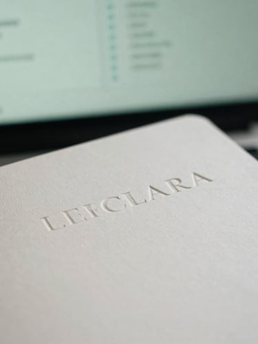 Close-up of an embossed LECLARA brand asset (e.g., stationery) on a clean desktop, with a blurred analytical dashboard in the background, natural overcast London daylight, soft shadows, macro detail.