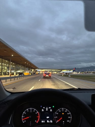 Driver-perspective shot from inside a vehicle approaching Málaga airport terminal access road at dusk, dashboard in sharp foreground silhouette, terminal lighting and aircraft tail visible through windshield, overcast sky, wide-angle framing conveying purposeful transit