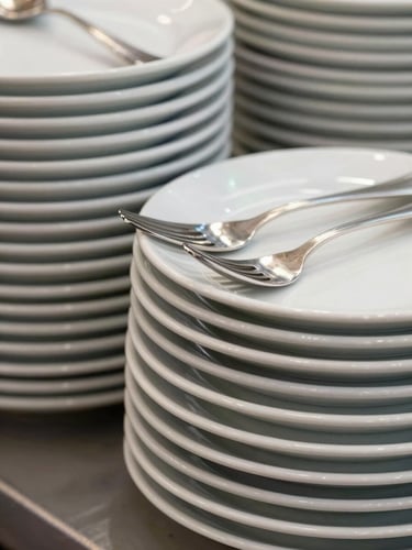 A close-up architectural shot of neatly stacked, spotless white ceramic plates and polished silver cutlery on a neutral surface
