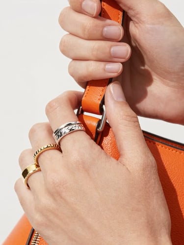 Macro close-up of dynamic hands wearing multiple textured silver and gold rings, holding a vibrant orange leather bag, bright high-key fashion photography, crisp natural daylight, sharp macro details, 35mm lens