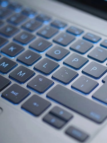 Close-up macro shot of a modern laptop keyboard, cool blue backlighting, sharp focus on the keys, subtle digital shield overlay, portrait aspect