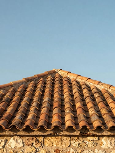 Low-angle shot looking up at a restored stone bastide roof with warm terracotta tiles against a clear blue sky, soft golden hour light, 35mm