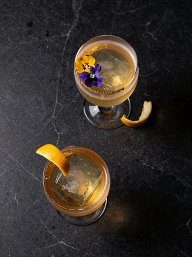 Overhead flat-lay of two handcrafted cocktails on a dark marble surface, one with a champagne gold rim and edible flower garnish, amber liquid catching warm back-light, a thin curl of citrus peel, negative dark space surrounding both glasses