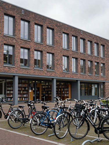 A clean, modern Dutch university campus library with brick architecture, large windows letting in bright overcast northern European daylight, and rows of bicycle racks in the foreground.