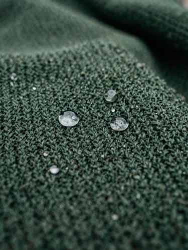 Macro close-up of water droplets beading on a dark green waterproof knitted blanket, overcast natural daylight, rich damp earth tones, sharp texture detail, 35mm