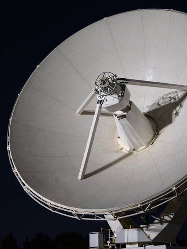 Close-up of a large satellite dish array at night, cool studio floodlights illuminating the dish surface from below, dense technical detail visible on the receiver housing, dark sky behind — shot tight on the dish face filling the frame edge to edge