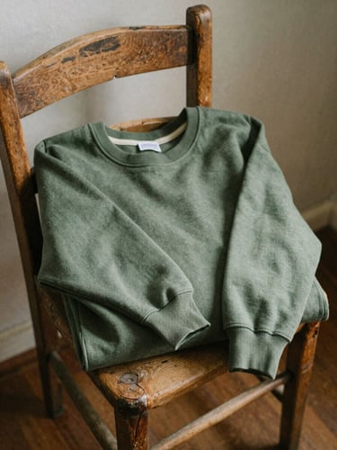 A heavy-weight olive green cotton sweatshirt folded on a rustic wooden chair in a sunlit cabin, natural ambient light, 35mm film grain