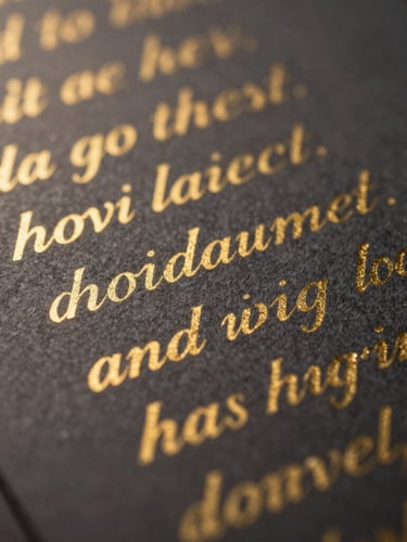 Extreme close-up macro shot of a handwritten lyric sheet on heavy, textured charcoal paper, gold ink calligraphy catching low-key dramatic light with warm gold backlights.