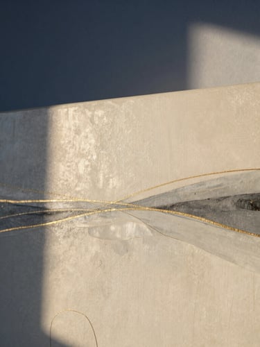 Wide shot of an abstract painting with layers of ivory, charcoal gray, and delicate gold lines, soft morning light