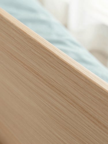Tight macro shot of light ash wood grain, showing a smooth, polished surface, with a hint of pastel blue fabric in the background, soft diffused morning light.