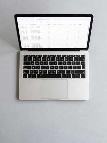 Overhead flat-lay of an organized desk with a sleek laptop displaying a clean spreadsheet, neutral gray surface, bright even office daylight.