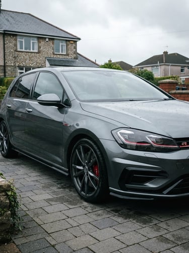 Low-angle three-quarter shot of a clean metallic grey German hatchback parked on a quiet Welsh suburban driveway, overcast soft daylight highlighting the sharp lines.