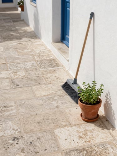Close-up overhead shot of a freshly swept Mediterranean courtyard, stone tiles clean and dry, a broom leaning against a whitewashed wall to the right, a small terracotta pot of herbs beside it, soft north-facing daylight