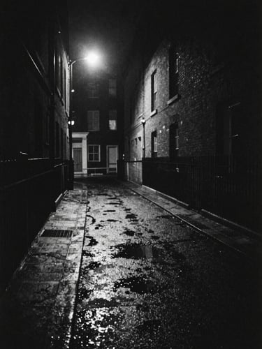 Wide atmospheric environmental shot of a dark London alleyway at night, harsh single streetlight casting long stark shadows on wet pavement, heavy film grain, gritty black and white