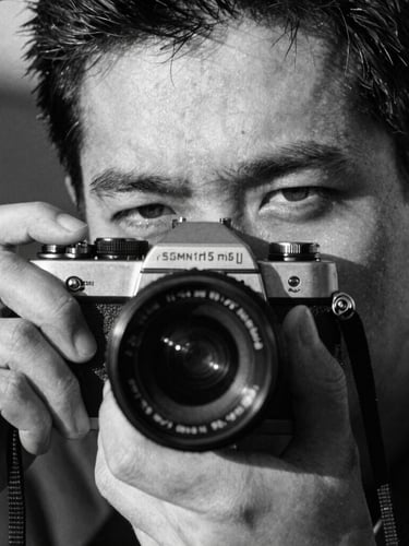 Extreme close-up portrait of a photographer holding a 35mm camera, harsh direct sunlight casting deep shadows across half the face, heavy film grain, gritty black and white photography, 35mm tri-x film style