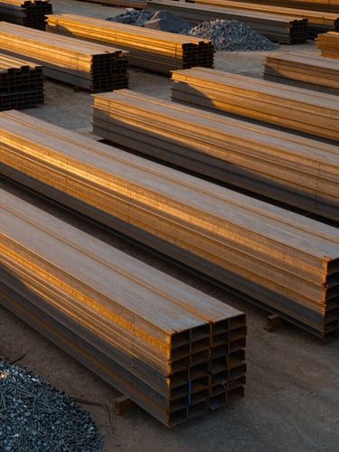 Wide shot of a material stockyard at golden hour — stacked steel beams and construction aggregate in organized rows, warm amber sunlight raking across the surfaces from the right, no people, industrial scale evident