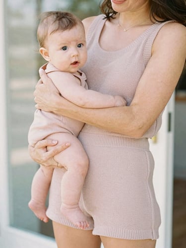 Candid lifestyle photo of a mother holding her baby wearing a soft dusty rose knit romper, gentle natural daylight, warm boutique aesthetic.
