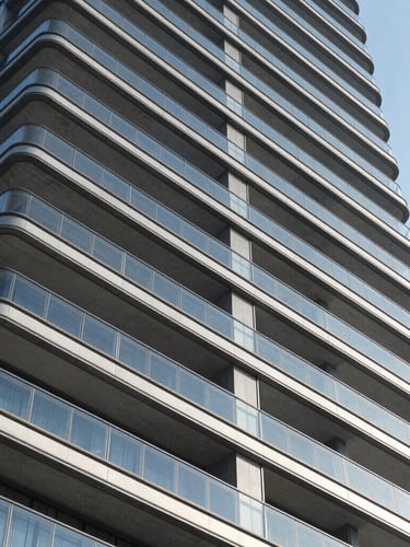 Low-angle architectural shot of a modern luxury residential tower facade, glass balconies reflecting blue sky, concrete geometric lines