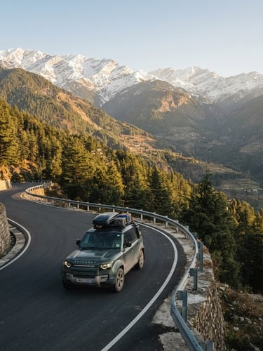 Wide environmental landscape of a private comfortable SUV driving on a clean winding mountain road in Shimla, surrounded by majestic snow-capped peaks and tall pine trees under warm morning golden sun.
