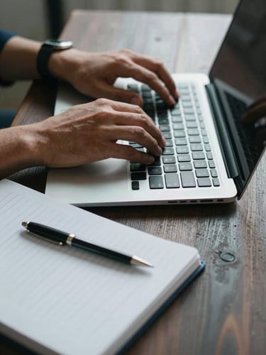 Close-up of hands typing on a laptop next to a structured notebook on a dark wood desk, cool daylight.