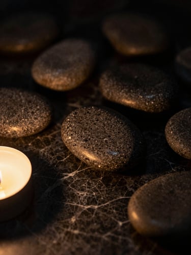 A macro close-up of smooth warm basalt stones resting on dark textured marble, soft golden candlelight casting deep shadows, moody editorial photography, 35mm