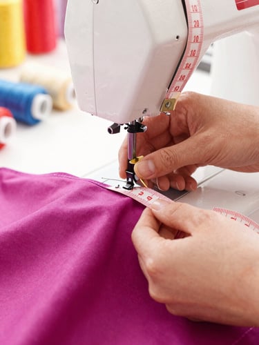 Close-up of hands carefully measuring a colorful magenta fabric with a soft measuring tape, sewing machine in a warm sunlit workshop, pet accessories visible in the background, shallow depth of field, 50mm